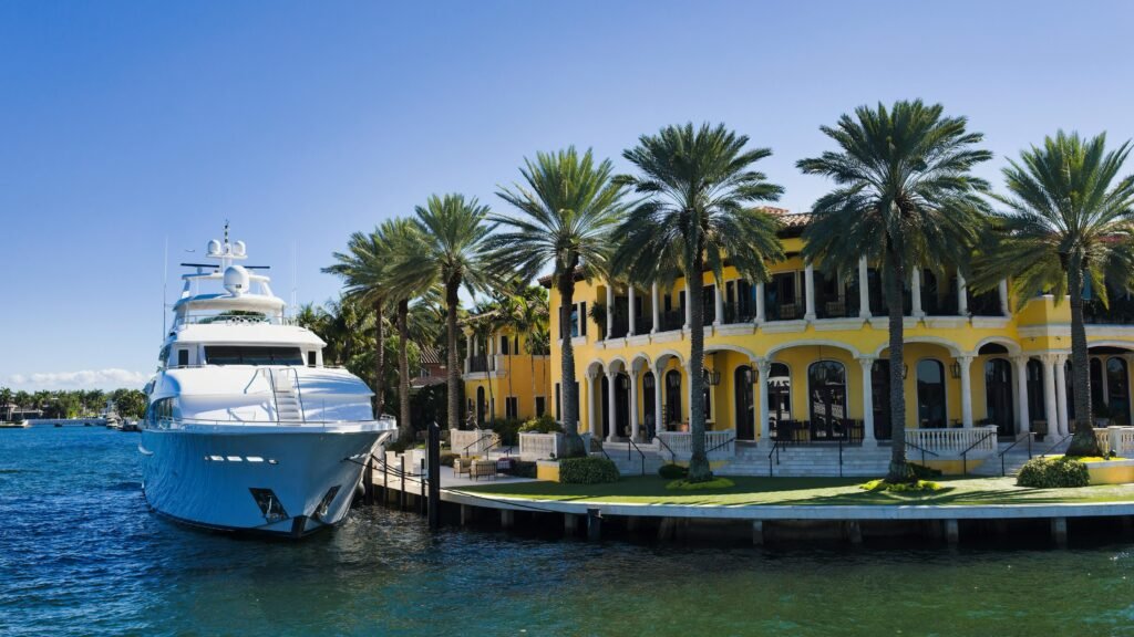 Luxury yacht docked beside a vibrant yellow waterfront mansion surrounded by palm trees in South Florida, representing upscale living and luxury home construction.